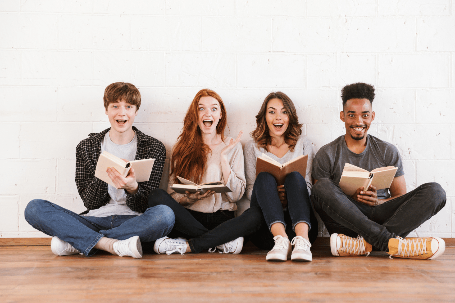 Four smiling college students sitting together with open books against a white brick wall